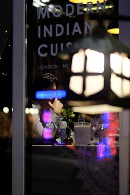 A person sitting at a table inside a restaurant, viewed through a window with reflections of neon lights. On the window, the words 'Modern Indian Cuisine' along with location names like 'London' and 'New' are visible. The table is set with a small vase containing a plant, a bottle of water, a glass, and a napkin holder. Dim, ambient lighting suggests a cozy dining atmosphere.