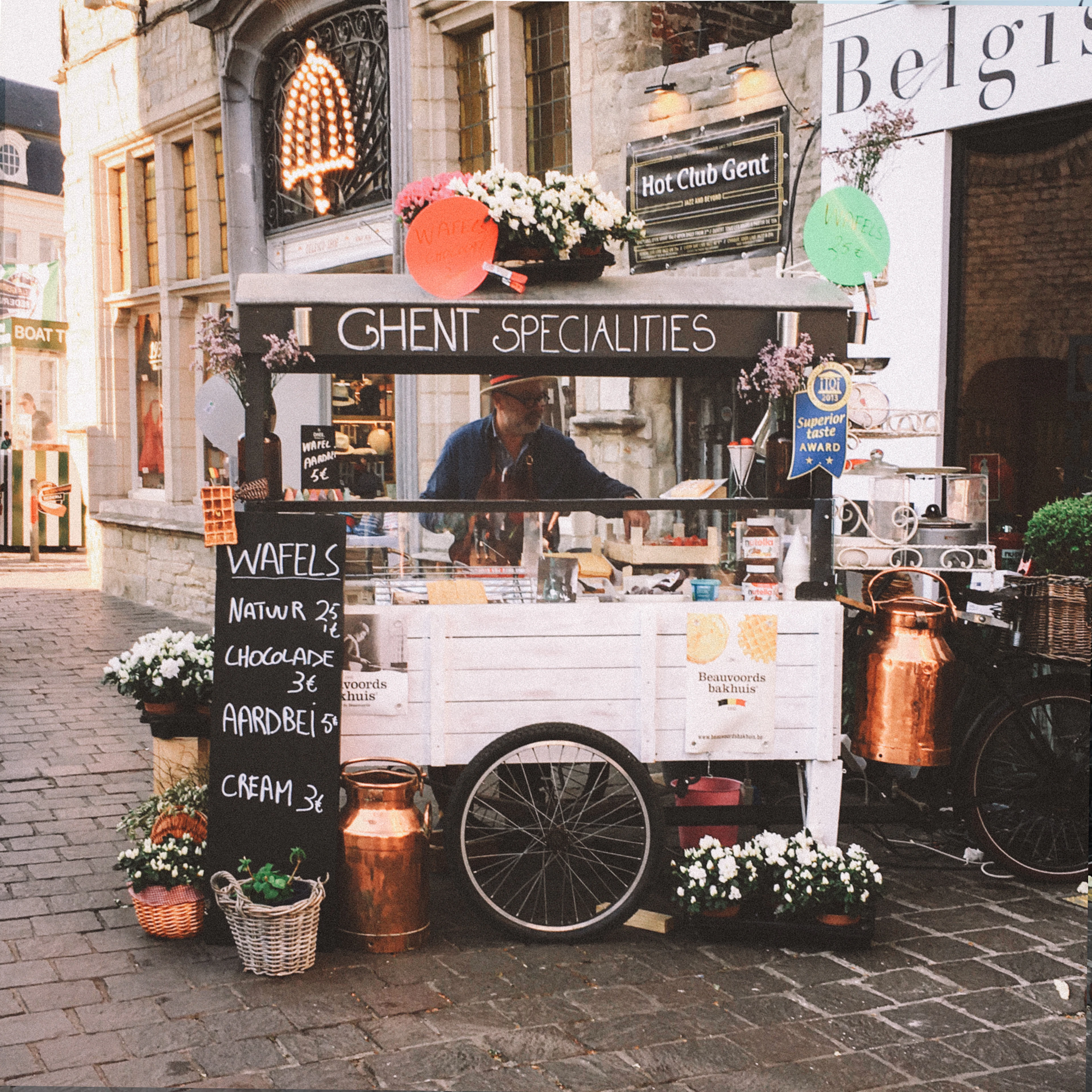 A waffle cart chalked "Ghent Specialities", banked with flowers on the cobbles