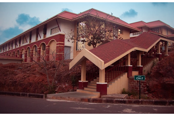 A campus building with red and cream brick architecture features a covered staircase. It is surrounded by earthy terrain and sparse vegetation. A sign indicating 'Hostel CH-7' is present near the stairs. The sky is slightly overcast, adding a serene atmosphere.