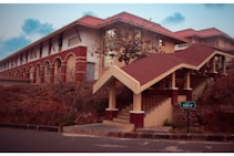A campus building with red and cream brick architecture features a covered staircase. It is surrounded by earthy terrain and sparse vegetation. A sign indicating 'Hostel CH-7' is present near the stairs. The sky is slightly overcast, adding a serene atmosphere.