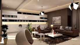A modern living room with contemporary design elements. The space features a large brown sectional sofa with patterned cushions, a sleek white coffee table, and a plush dark brown rug. Stylish pendant lights hang from the ceiling, and a monochrome artwork adorns one of the walls. Books and decorative items are neatly arranged in a long shelf in the background. The room is accentuated by potted plants and a uniquely shaped modern chair.