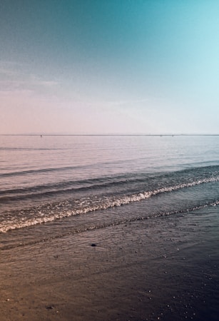 Calm ocean waves gently lap at the sandy shore under a clear sky. The horizon is faintly visible in the distance, separating the vast expanse of sea and sky. The scene conveys a sense of peace and tranquility.
