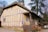 A charming, rustic cottage features a thatched roof and timber-framed walls with a yellowish hue. It is surrounded by large trees and has a front porch lined with various clay pots. There are outdoor chairs and a table nearby, giving it a cozy, pastoral feel.