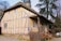 A charming, rustic cottage features a thatched roof and timber-framed walls with a yellowish hue. It is surrounded by large trees and has a front porch lined with various clay pots. There are outdoor chairs and a table nearby, giving it a cozy, pastoral feel.