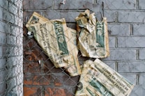 A crumpled newspaper caught in a chain-link fence against a brick wall. The newspaper is positioned haphazardly and shows signs of wear and tear.