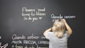 A person is standing in front of a large chalkboard, writing motivational phrases in different colors. The board has phrases in both English and Portuguese. The person has short, light-colored hair and is wearing a gray shirt.