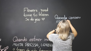 A person is standing in front of a large chalkboard, writing motivational phrases in different colors. The board has phrases in both English and Portuguese. The person has short, light-colored hair and is wearing a gray shirt.
