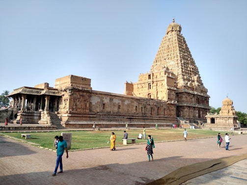 An ancient temple displaying intricate architecture with a large tower, surrounded by several smaller structures and lush greenery. People are walking and standing in front of the temple, capturing the essence of historical and cultural ambiance.