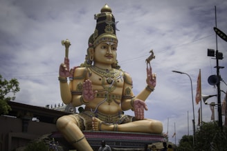 A large statue of a deity adorned with intricate jewelry and traditional attire, seated in a cross-legged position. The statue holds a small deer in one hand and a trident in another, surrounded by a cloudy sky and various objects in the background.
