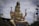 A large statue of a deity adorned with intricate jewelry and traditional attire, seated in a cross-legged position. The statue holds a small deer in one hand and a trident in another, surrounded by a cloudy sky and various objects in the background.