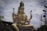A large statue of a deity adorned with intricate jewelry and traditional attire, seated in a cross-legged position. The statue holds a small deer in one hand and a trident in another, surrounded by a cloudy sky and various objects in the background.