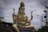 A large statue of a deity adorned with intricate jewelry and traditional attire, seated in a cross-legged position. The statue holds a small deer in one hand and a trident in another, surrounded by a cloudy sky and various objects in the background.