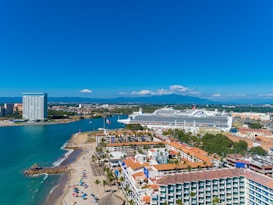 A coastal cityscape featuring a large cruise ship docked near a bustling port area. The foreground includes a beach with colorful umbrellas and palm trees. A prominent modern high-rise building is visible next to the water, along with a wide view of the cityscape and distant mountains under a clear blue sky.
