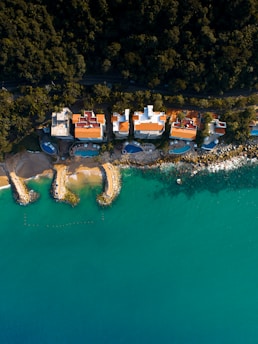 An aerial view of a coastal area with several houses lined along the edge of a lush green forest. The houses feature bright red and orange roofs and are adjacent to a calm, turquoise sea. Several rock formations extend into the water, forming small bays with sandy beaches.