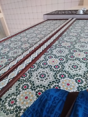Intricate geometric tilework featuring a complex pattern of stars and polygons with colors like green, red, white, and yellow. The pattern repeats across the surface, bordered by wooden strips. A portion of a tiled wall and a blue fabric with text can also be seen.