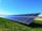 A solar panel array is installed on a grassy field under a clear blue sky. The panels are angled to capture maximum sunlight, and they are placed in a line stretching into the distance.