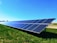 A solar panel array is installed on a grassy field under a clear blue sky. The panels are angled to capture maximum sunlight, and they are placed in a line stretching into the distance.
