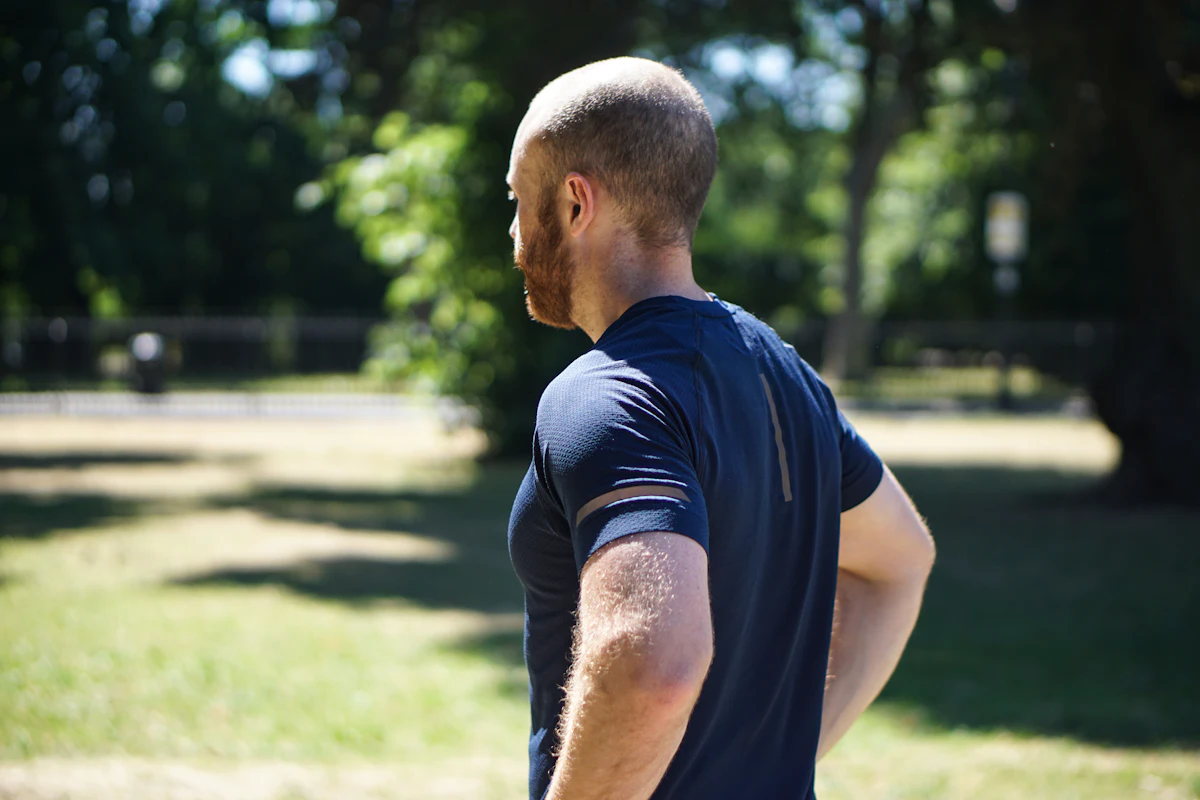 Pelari pria berdiri dengan tangan di pinggang, diambil dari belakang; jersey training navy dengan strip reflektif di lengan, latar tangan saat siang hari