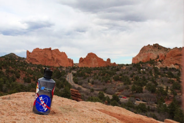 A bottle with a NASA logo rests on a rock in the foreground, set against a backdrop of jagged red rock formations and a winding road. The scene is surrounded by greenery and under a cloudy sky.