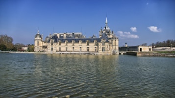 A grand, historic chateau with intricate architectural details is situated beside a large body of water, with a bright blue sky and a few fluffy clouds in the background. The landscape features surrounding trees, adding to the serene and elegant atmosphere.