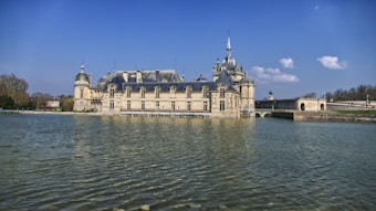 A grand, historic chateau with intricate architectural details is situated beside a large body of water, with a bright blue sky and a few fluffy clouds in the background. The landscape features surrounding trees, adding to the serene and elegant atmosphere.