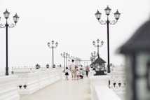 A group of people is walking and interacting on a long, elegant pier adorned with ornate black street lamps. The pier features intricate white railings and lamp posts, creating a classic and serene atmosphere. The scene is open and airy, and the background appears misty, blending with the light colors of the sky.