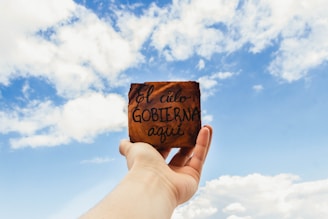 A hand holds up a small wooden plaque with the handwritten words 'El cielo GOBIERNA aquí' against a backdrop of a bright blue sky with scattered white clouds.