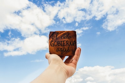 A hand holds up a small wooden plaque with the handwritten words 'El cielo GOBIERNA aquí' against a backdrop of a bright blue sky with scattered white clouds.