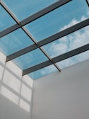 A modern architectural ceiling design featuring a glass roof, through which a clear blue sky with some clouds is visible. Metal beams create a grid pattern across the glass panels, casting geometric shadows on the adjacent white wall.