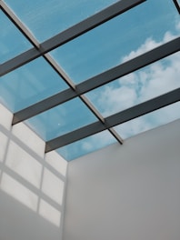 A modern architectural ceiling design featuring a glass roof, through which a clear blue sky with some clouds is visible. Metal beams create a grid pattern across the glass panels, casting geometric shadows on the adjacent white wall.