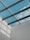 A modern architectural ceiling design featuring a glass roof, through which a clear blue sky with some clouds is visible. Metal beams create a grid pattern across the glass panels, casting geometric shadows on the adjacent white wall.
