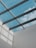 A modern architectural ceiling design featuring a glass roof, through which a clear blue sky with some clouds is visible. Metal beams create a grid pattern across the glass panels, casting geometric shadows on the adjacent white wall.