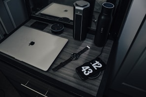 A workspace with a laptop, smart speaker, smartwatch, and a phone displaying a clock, placed on a wooden desk. The setup includes a water bottle and another cylindrical object, reflecting a modern and minimalist style.