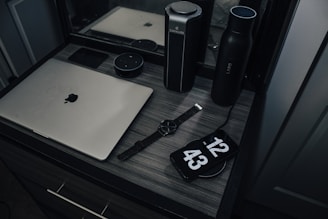 A workspace with a laptop, smart speaker, smartwatch, and a phone displaying a clock, placed on a wooden desk. The setup includes a water bottle and another cylindrical object, reflecting a modern and minimalist style.
