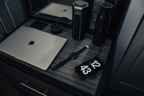 A workspace with a laptop, smart speaker, smartwatch, and a phone displaying a clock, placed on a wooden desk. The setup includes a water bottle and another cylindrical object, reflecting a modern and minimalist style.