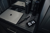 A workspace with a laptop, smart speaker, smartwatch, and a phone displaying a clock, placed on a wooden desk. The setup includes a water bottle and another cylindrical object, reflecting a modern and minimalist style.