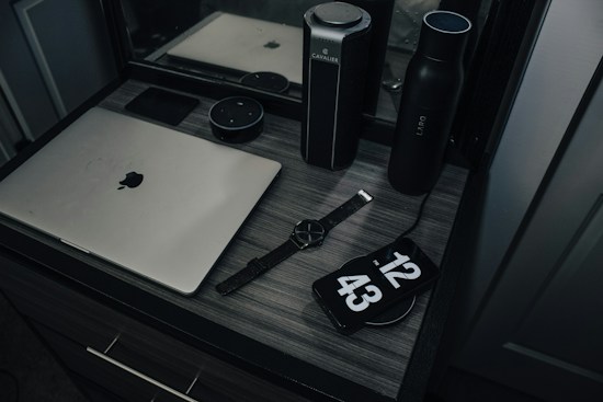 A workspace with a laptop, smart speaker, smartwatch, and a phone displaying a clock, placed on a wooden desk. The setup includes a water bottle and another cylindrical object, reflecting a modern and minimalist style.