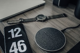 A wooden desk features several electronic items including a smartwatch with a black strap, a smartphone displaying the time '12:46 PM' in large digital numbers, and a circular wireless charging pad labeled 'FAST CHARGE'. A laptop is partially visible in the top left corner of the image.