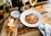 A plate of penne pasta topped with grated cheese and herbs is placed on a wooden table. Nearby, there is a wooden board with freshly sliced bread and a small bowl with grated cheese. A bottle of wine and a glass of white wine are also present. On the side, there is a bottle of olive oil with a vine-like design. Forks and a napkin are positioned to the right.