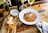A plate of penne pasta topped with grated cheese and herbs is placed on a wooden table. Nearby, there is a wooden board with freshly sliced bread and a small bowl with grated cheese. A bottle of wine and a glass of white wine are also present. On the side, there is a bottle of olive oil with a vine-like design. Forks and a napkin are positioned to the right.