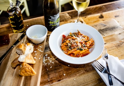 A plate of penne pasta topped with grated cheese and herbs is placed on a wooden table. Nearby, there is a wooden board with freshly sliced bread and a small bowl with grated cheese. A bottle of wine and a glass of white wine are also present. On the side, there is a bottle of olive oil with a vine-like design. Forks and a napkin are positioned to the right.