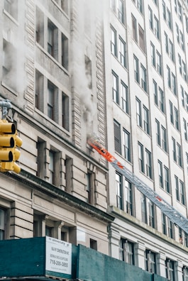 Smoke billows from a window on an upper floor of a tall building, with a fire truck's ladder extended towards the window. The facade of the building shows intricate architectural details. A yellow traffic light is visible in the foreground, along with construction scaffolding at the street level.