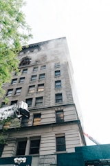 A multi-story building with classic architectural details shows smoke emanating from several windows. Near the bottom, construction equipment and scaffolding are visible, and green tree branches partially frame the view.