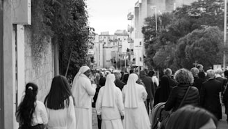 A group of people, including several wearing religious habits, walk down a street lined with buildings and trees. The scene suggests a communal or procession-like activity in an urban setting.