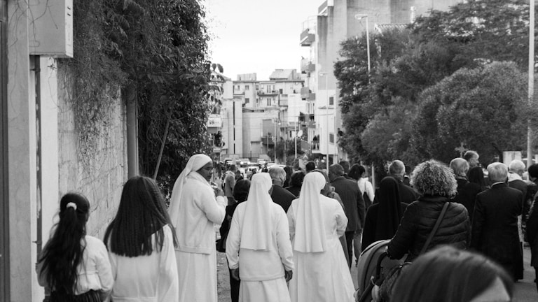 A group of people, including several wearing religious habits, walk down a street lined with buildings and trees. The scene suggests a communal or procession-like activity in an urban setting.