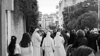 A group of people, including several wearing religious habits, walk down a street lined with buildings and trees. The scene suggests a communal or procession-like activity in an urban setting.