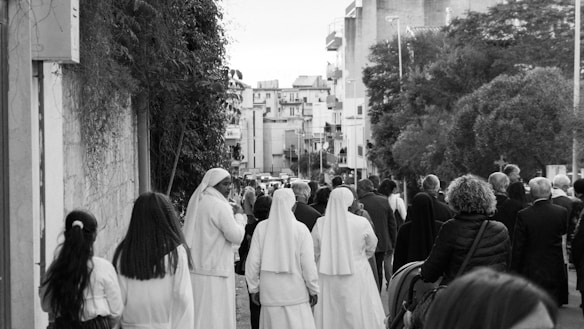 A group of people, including several wearing religious habits, walk down a street lined with buildings and trees. The scene suggests a communal or procession-like activity in an urban setting.