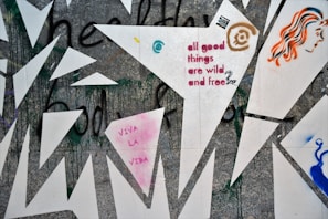 A graffiti-covered wall features geometric shapes and vibrant colors. Among the designs are the phrases 'all good things are wild and free' and 'VIVA LA VIDA'. A stylized face with orange and blue hair and abstract patterns add to the complexity of the artwork.
