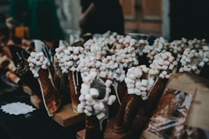 Waffle cones filled with small marshmallows are displayed on a table. The cones appear to be drizzled with chocolate or a similar topping, adding to their visual appeal. The setting seems to be a market or a stall.