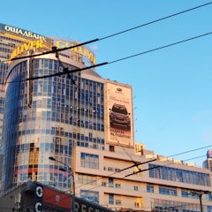 A modern, multi-story building with reflective glass windows featuring a large billboard advertisement for a car model. The building has the name 'Gulliver' prominently displayed in yellow on the roof. In the foreground, there are visible overhead power lines. The sky in the background is clear and blue.